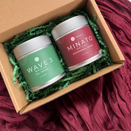 Two small containers labeled 'Wave 3' and 'Minato' in a cardboard box with green filler on a maroon fabric background.