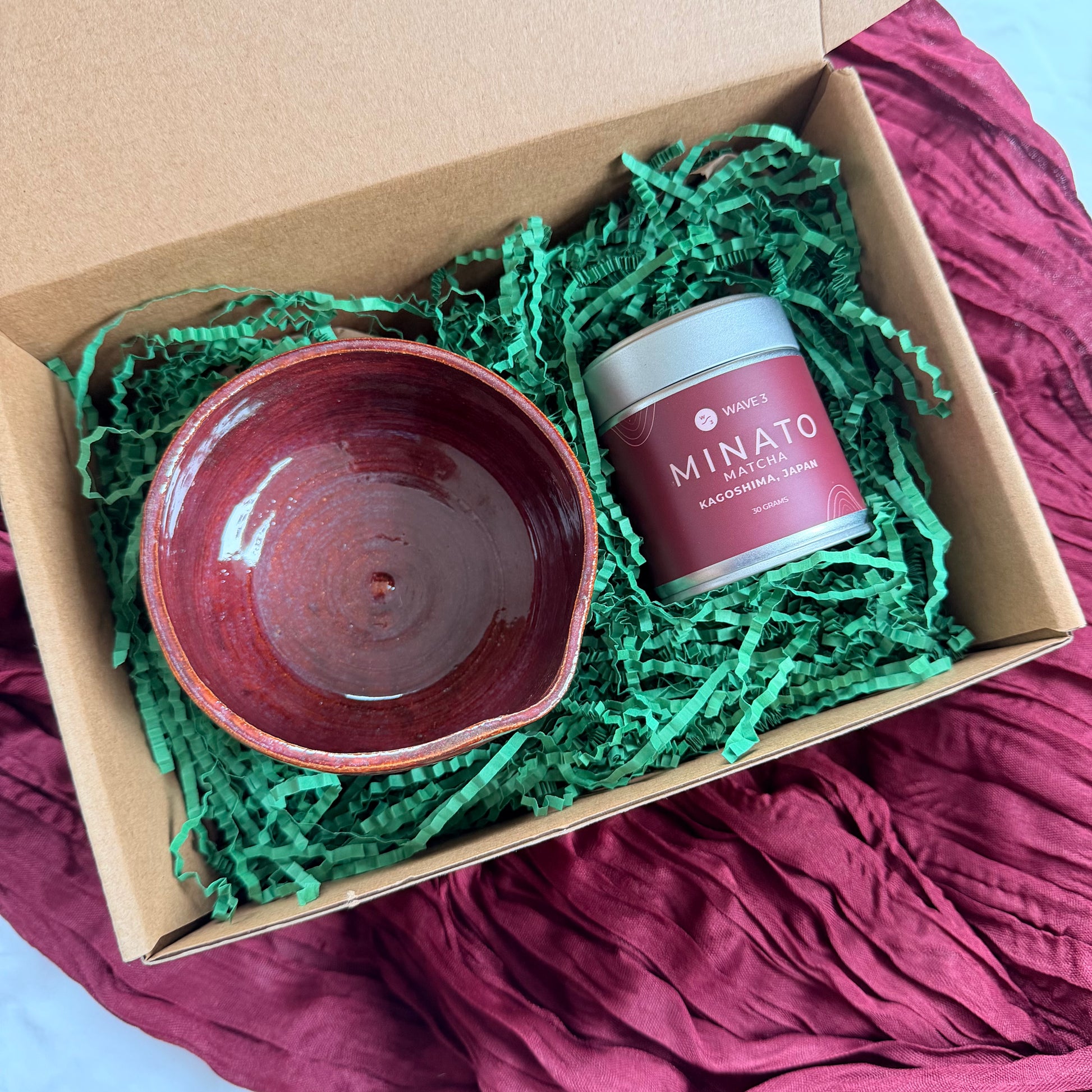 Red ceramic bowl and pink jar labeled 'MINATO' in a cardboard box with green shredded paper on a red fabric background.